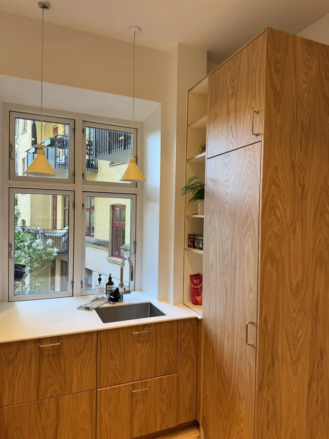 Køkken på Nørrebro efter renovering med egetræsskabe, hvid bordplade og gule pendler ved vinduet